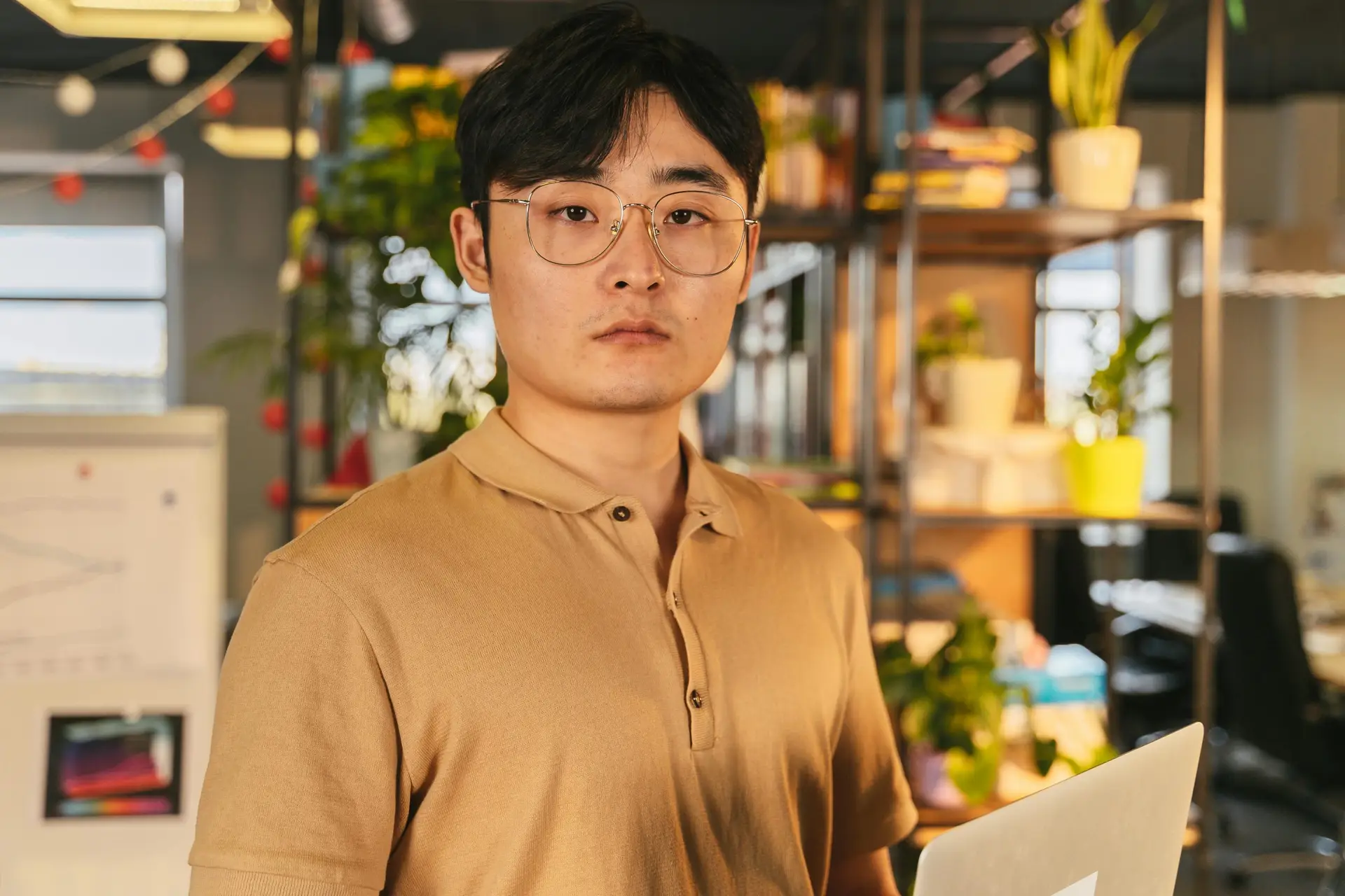 A serious Asian man wearing eyeglasses and a polo shirt, standing indoors with a laptop.